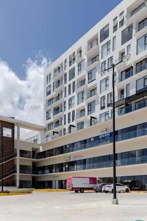 A multi-story hotel with a modern design, featuring balconies, large windows, and a parking area with vehicles. - Cancun 1BR | Lagoon View | Rooftop Pool & Gym (Cancún)