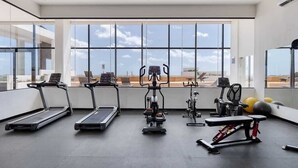 Et fitnesscenter med løbebånd, crosstrainer, bænkpres og træningsbolde, store vinduer og udsigt til himlen.
