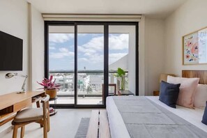 A modern bedroom with a large sliding glass door, a bed with pillows, a desk with a chair, and a potted plant. - Cancun 1BR | Lagoon View | Rooftop Pool & Gym (Cancún)