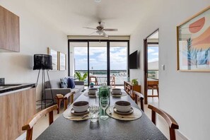 A modern dining area with a view of the sea, a ceiling fan, and a dining table set for four. - Cancun 1BR | Lagoon View | Rooftop Pool & Gym (Cancún)