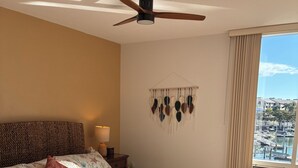 Quarto com uma cama grande, um ventilador de teto, uma decoração de macramé na parede e vista para uma marina através de uma janela.