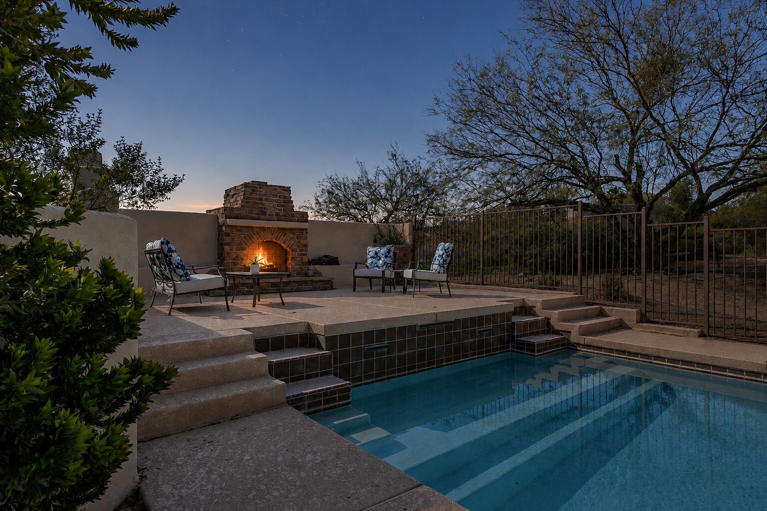 A pool with steps leading into it, surrounded by a patio area with chairs and a stone fireplace.