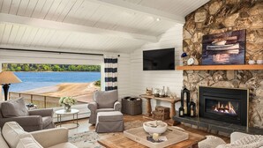 Ein gemütliches Wohnzimmer mit Kamin aus Stein, bequemen Sitzmöglichkeiten, einem Fernseher und Blick auf den Strand.