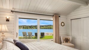 Ein Schlafzimmer mit einem großen Bett, Seeblick, zwei Stühlen draußen und einer Tür rechts.