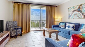 A living room with a sofa, armchair, coffee table, and a TV stand. There is a balcony with curtains and a view of the sky. - Lakefront condo with amazing views, pool, & hot tub - steps from the beach (Orlando)
