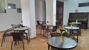 A dining area with round tables and wicker chairs, a piano, and a door leading to another room. - Flor y Canto  (Matehuala)
