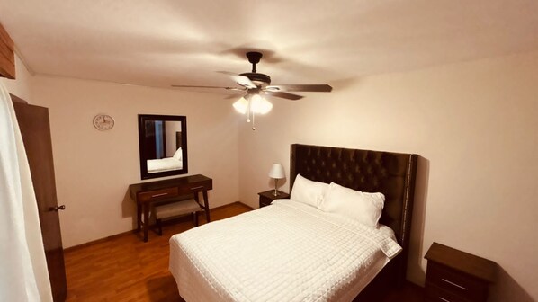 Un dormitorio con cama, escritorio, silla, mesita de noche, lámpara, ventilador de techo y un reloj en la pared.