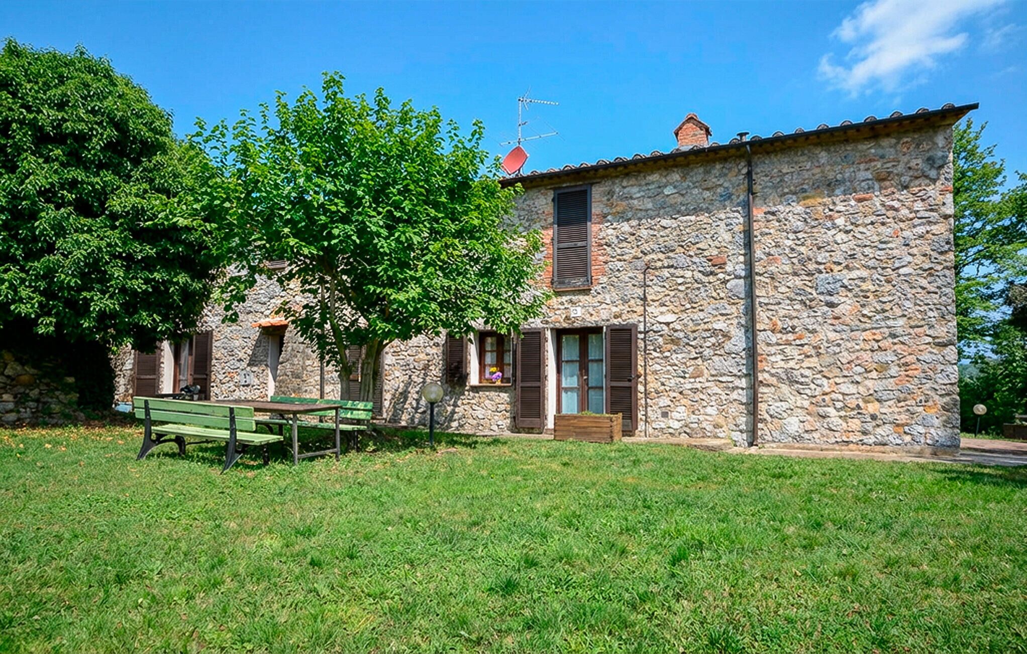A stone building with a wooden bench in front, a tree to the left, and a clear blue sky.