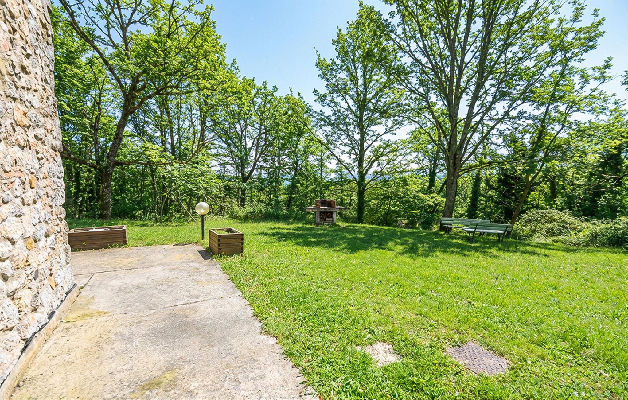 A grassy outdoor area with a stone wall, a wooden bench, and a table with a lamp.