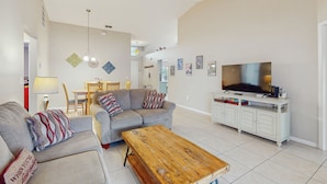 A living room with a grey sofa, a wooden coffee table, a TV on a white cabinet, and a dining area in the background. - Spacious Retreat with Private Pool, Enclosed Yard, WiFi & Game Room (Clermont)