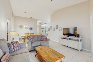 A living room with a grey sofa, a wooden coffee table, a TV on a white cabinet, and a dining area in the background. - Spacious Retreat with Private Pool, Enclosed Yard, WiFi & Game Room (Clermont)