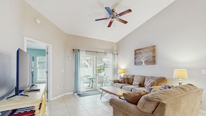 A living room with a television, sofa, and coffee table. There is a ceiling fan, a door leading to a balcony, and a painting on the wall. - Spacious Retreat with Private Pool, Enclosed Yard, WiFi & Game Room (Clermont)