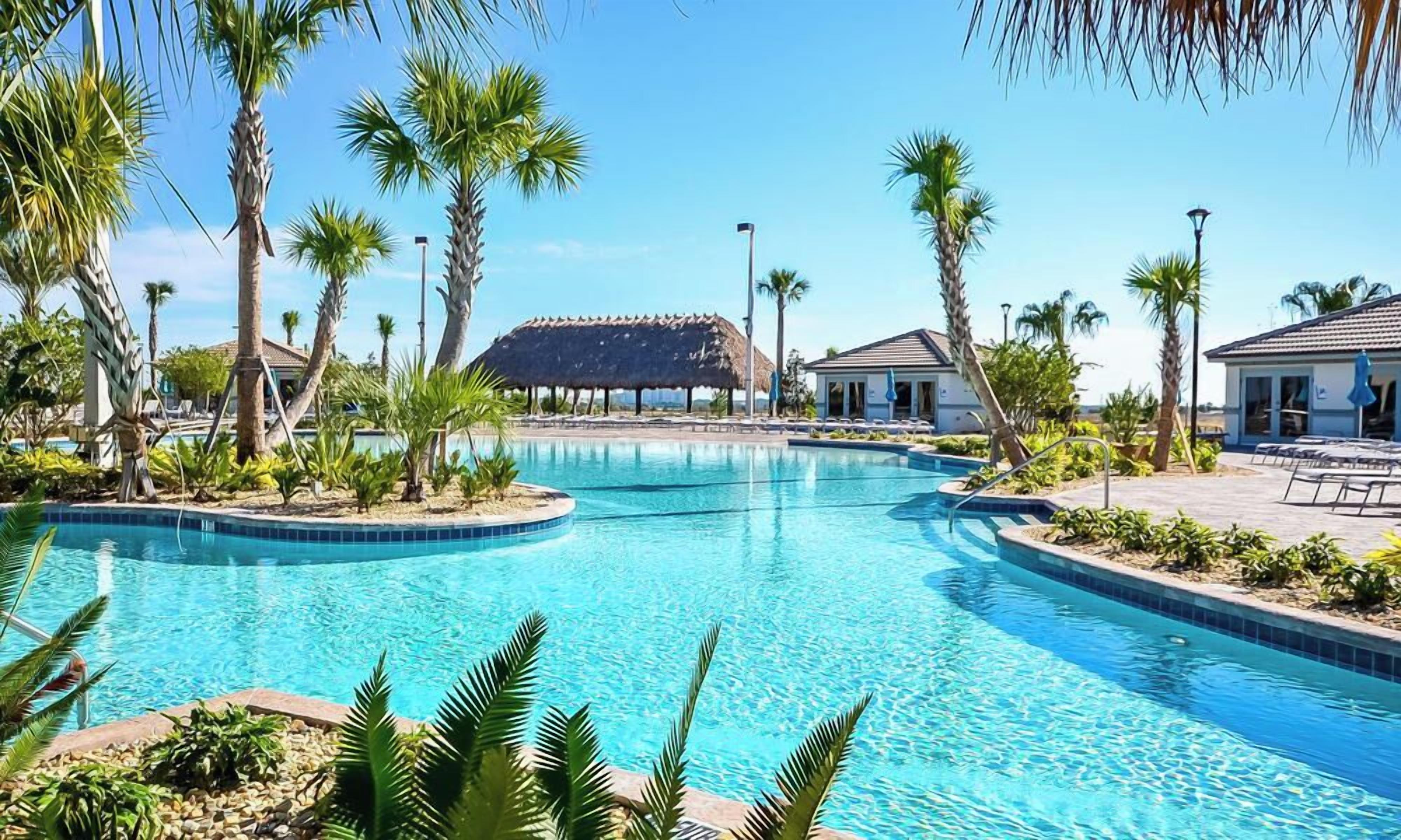 A swimming pool with a thatched-roof structure in the background, surrounded by palm trees and lounge chairs.