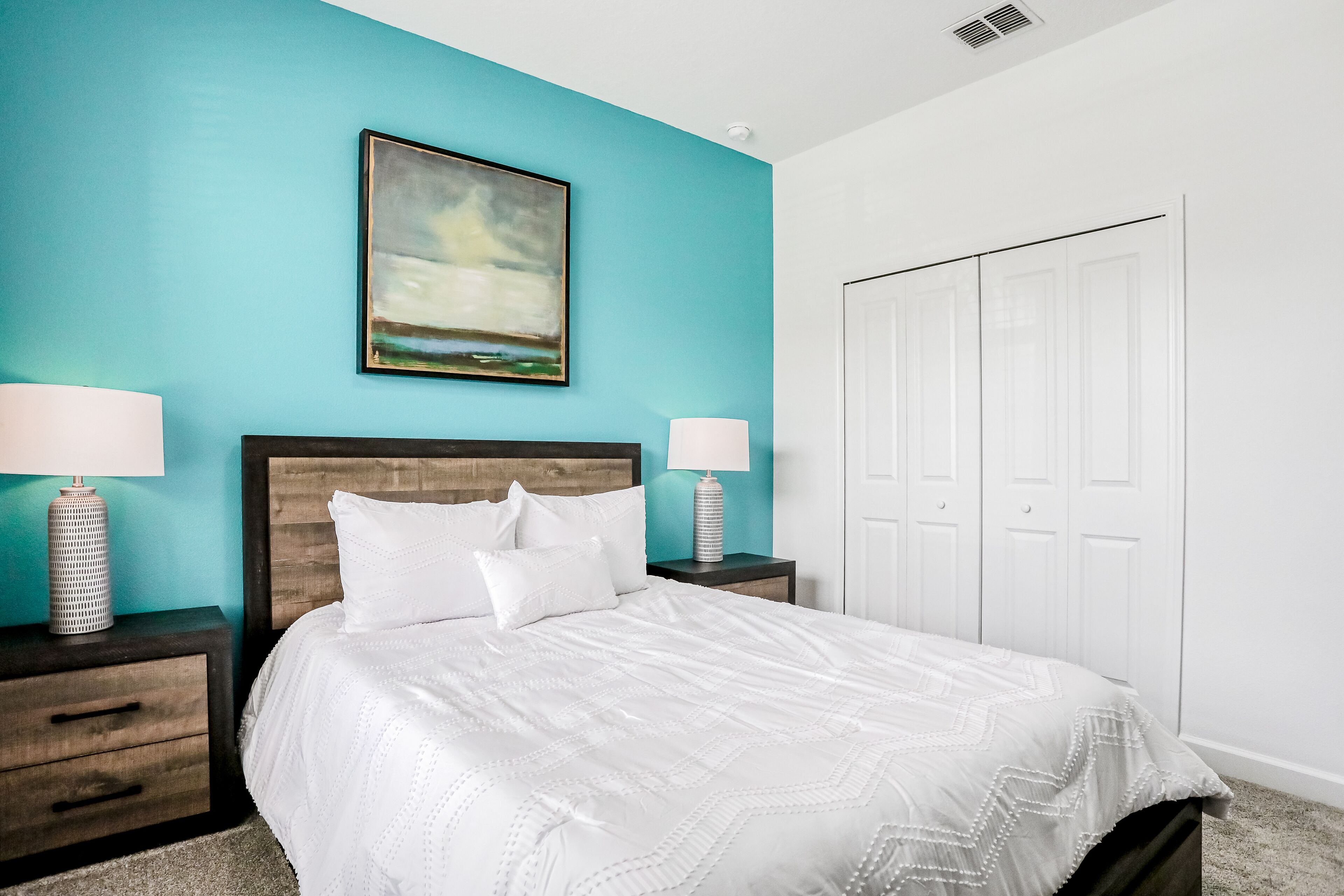 A bedroom with a bed, two bedside tables with lamps, a painting on the wall, and a white wardrobe.