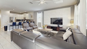 A modern living room with a grey sectional sofa, a coffee table, and a flat-screen TV. There is a kitchen area with white cabinets and a microwave.