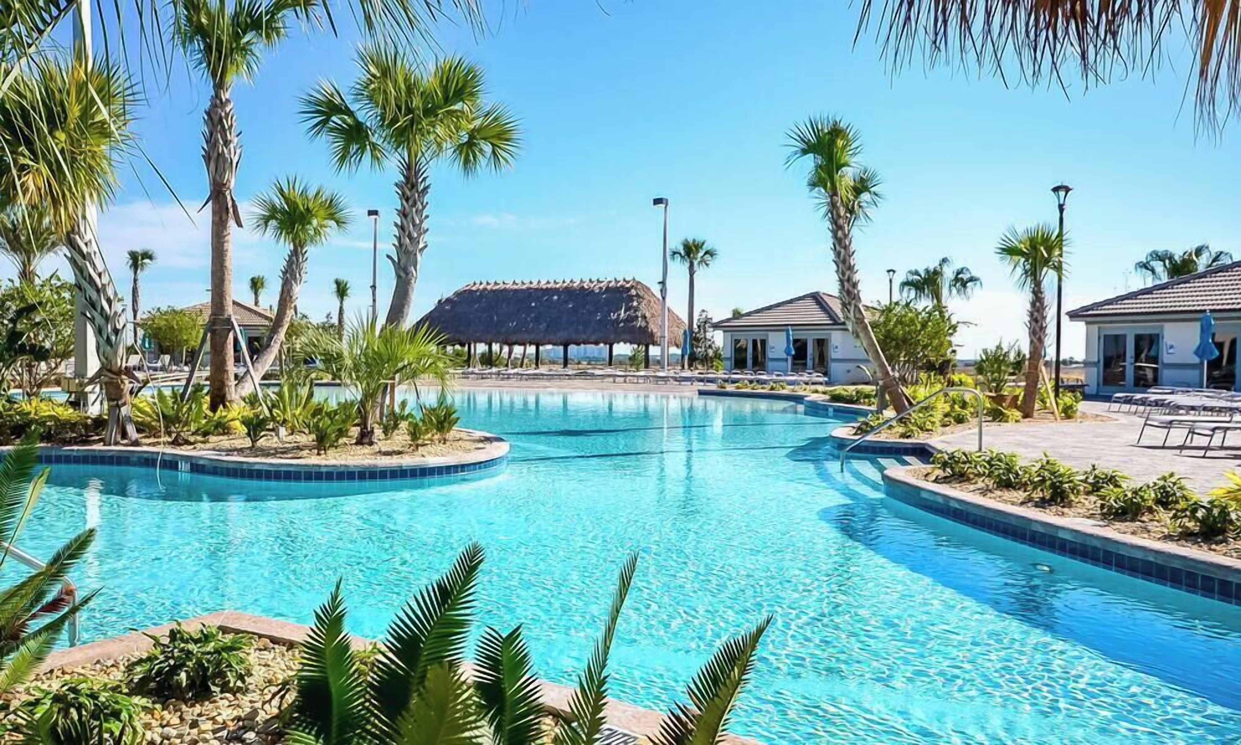 A swimming pool with a thatched-roof structure in the background, surrounded by palm trees and lounge chairs.