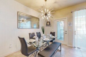 Un espace repas comprenant une table en verre, des chaises noires, un banc, un lustre, un grand miroir et une porte donnant sur l’extérieur.