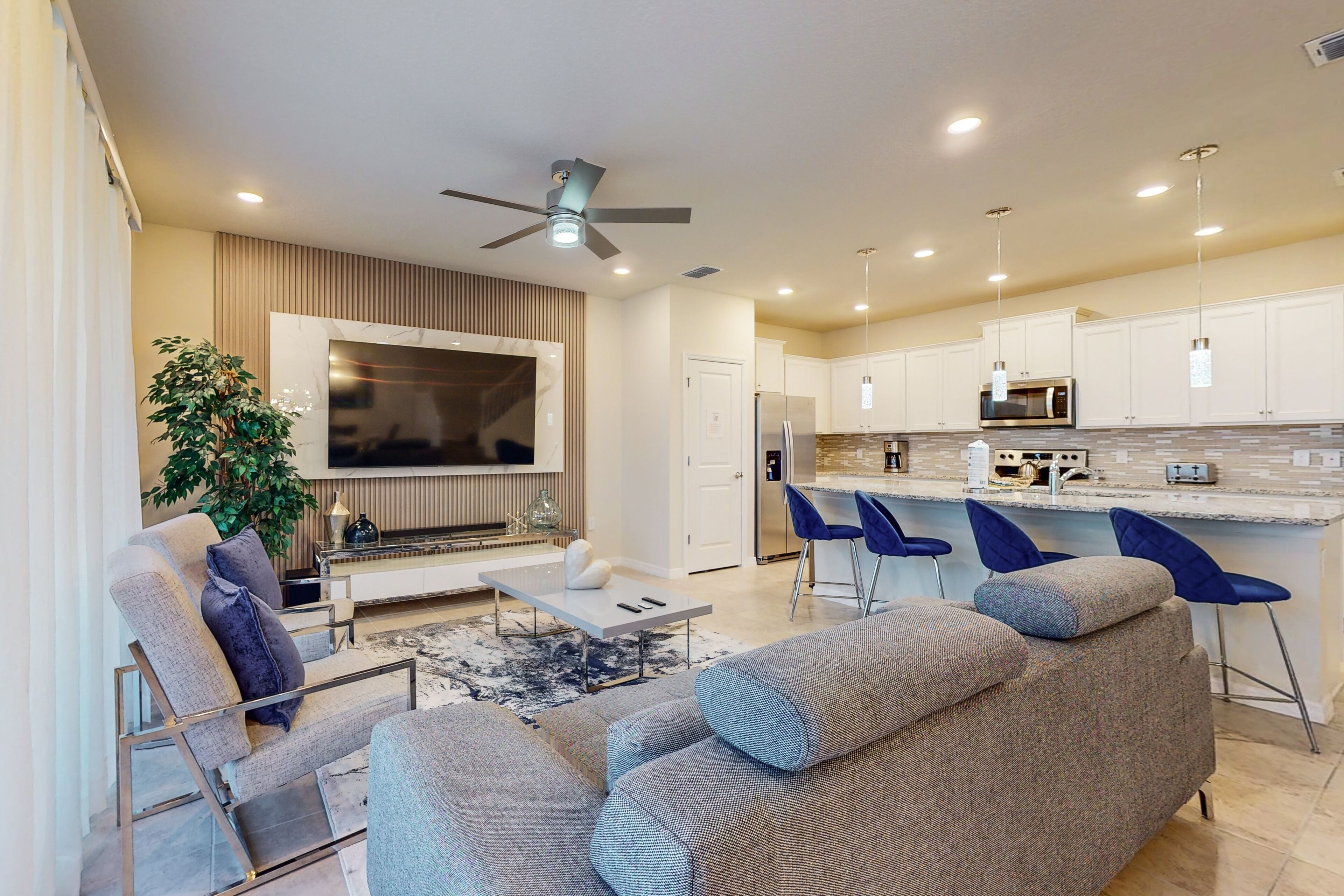 Una sala de estar moderna con sofá, una mesa de centro y televisor. Hay una zona de cocina con microondas, refrigerador y taburetes de bar.