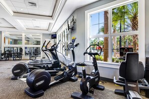 Ein Fitnessstudio mit Laufbändern, Crosstrainern und Kraftgeräten, großen Fenstern mit Blick auf Palmen und einer Decke mit eingelassener Beleuchtung.