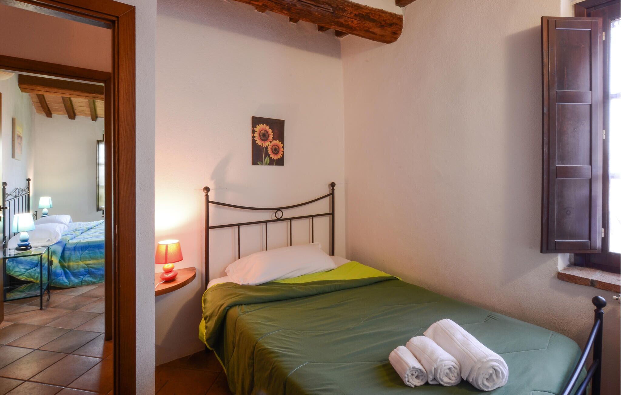 Una cama individual con colcha verde, una mesita de noche con lámpara, y un cuadro de girasoles en la pared.