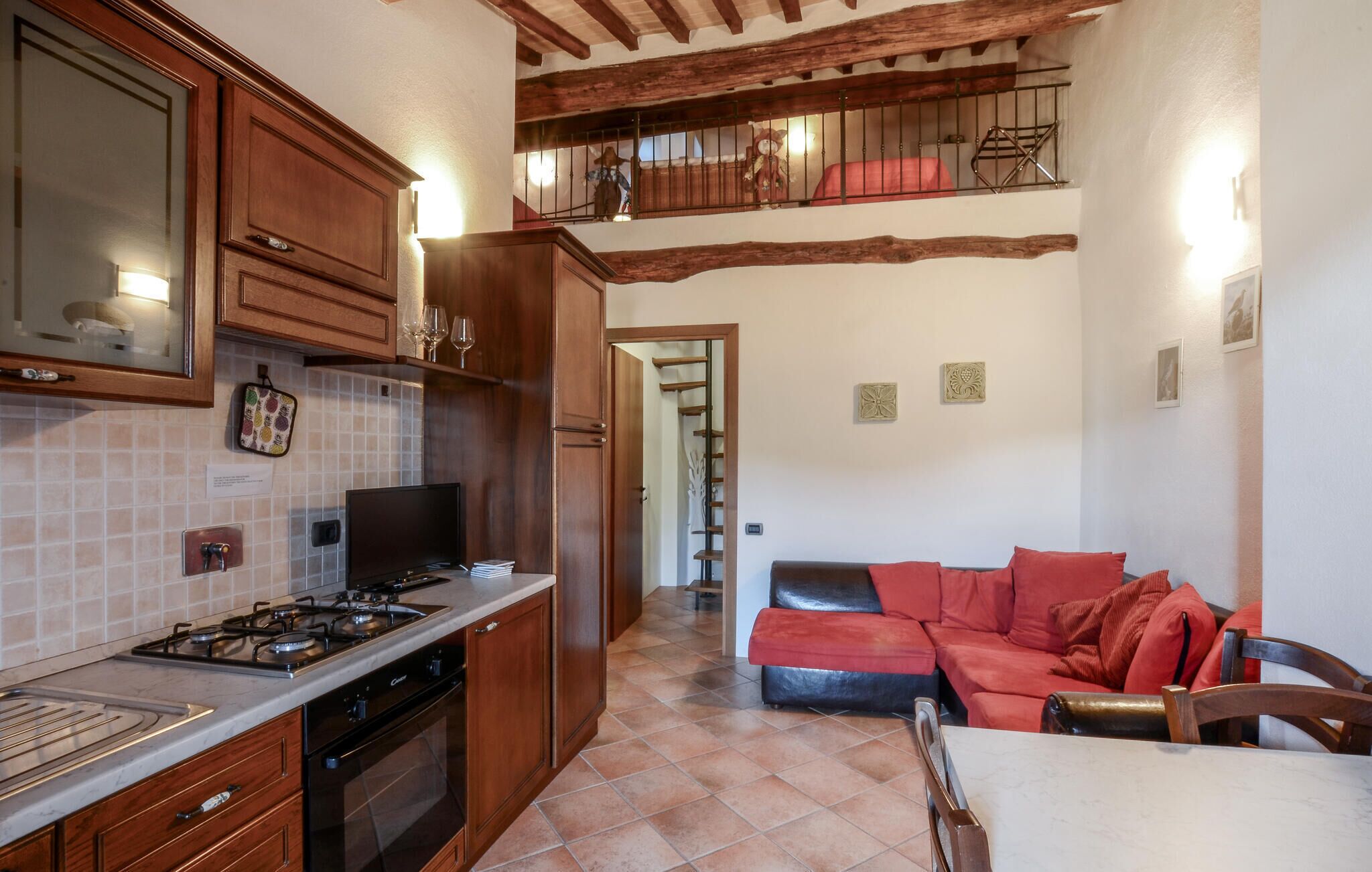 A kitchen with wooden cabinets, a gas stove, and a tiled backsplash. There is a dining area with a table and chairs, and a red sofa. A staircase leads to an upper level with a balcony.