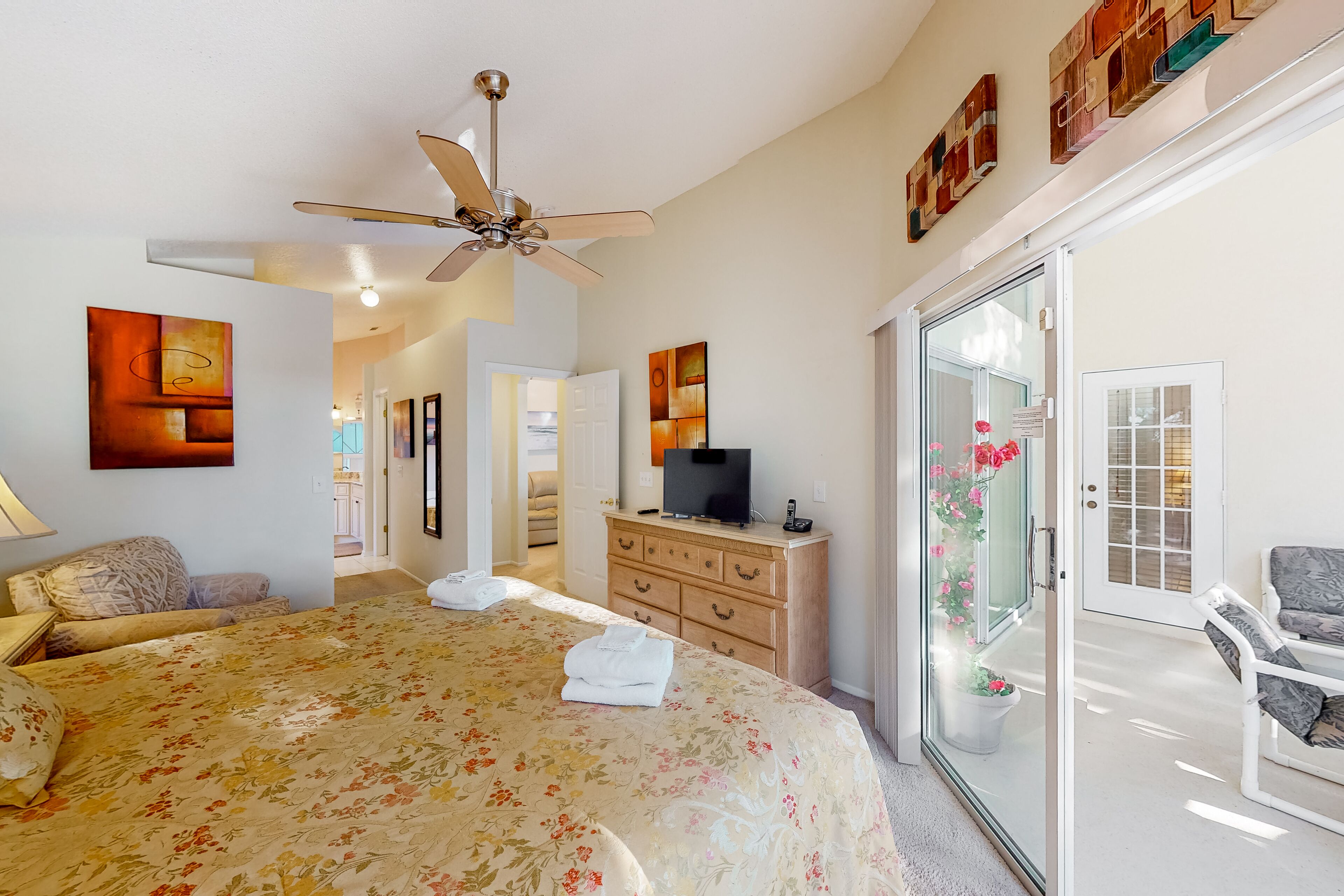 A bedroom with a bed, a ceiling fan, a dresser, a television, and a sliding glass door leading to a balcony with flowers.