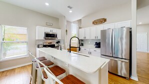 A modern kitchen with white cabinetry, a stainless steel refrigerator, a central island with stools, and a window with blinds. - Harmony Villa | Family-friendly home in Davenport with a game room & pool (Davenport)