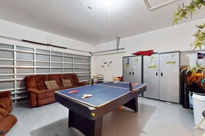 Une pièce avec une table de ping-pong, des canapés marron, un réfrigérateur et une étagère fixée au mur.