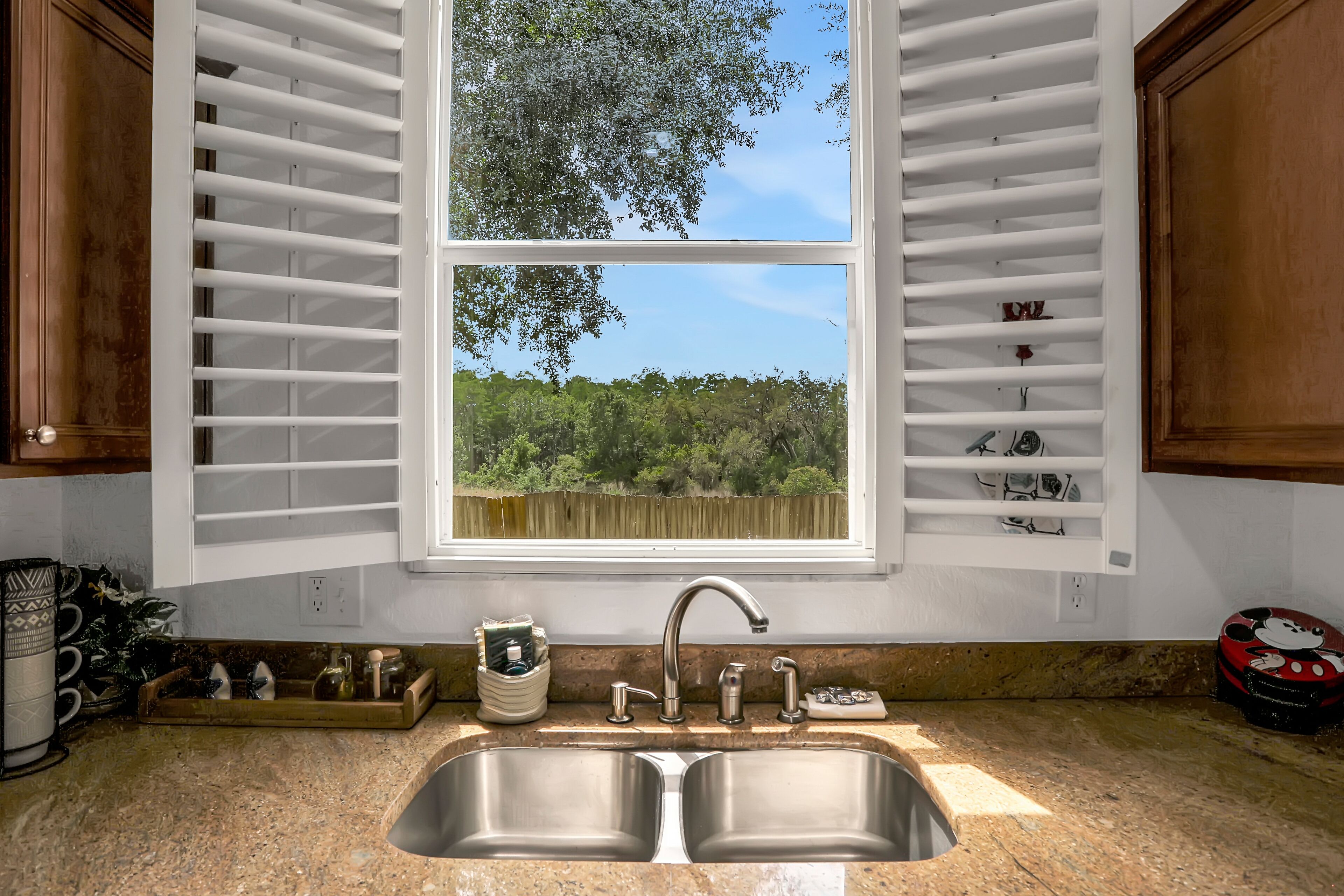 Eine Küche mit doppeltem Spülbecken, einem Fenster mit Jalousien und Blick auf einen Wald.