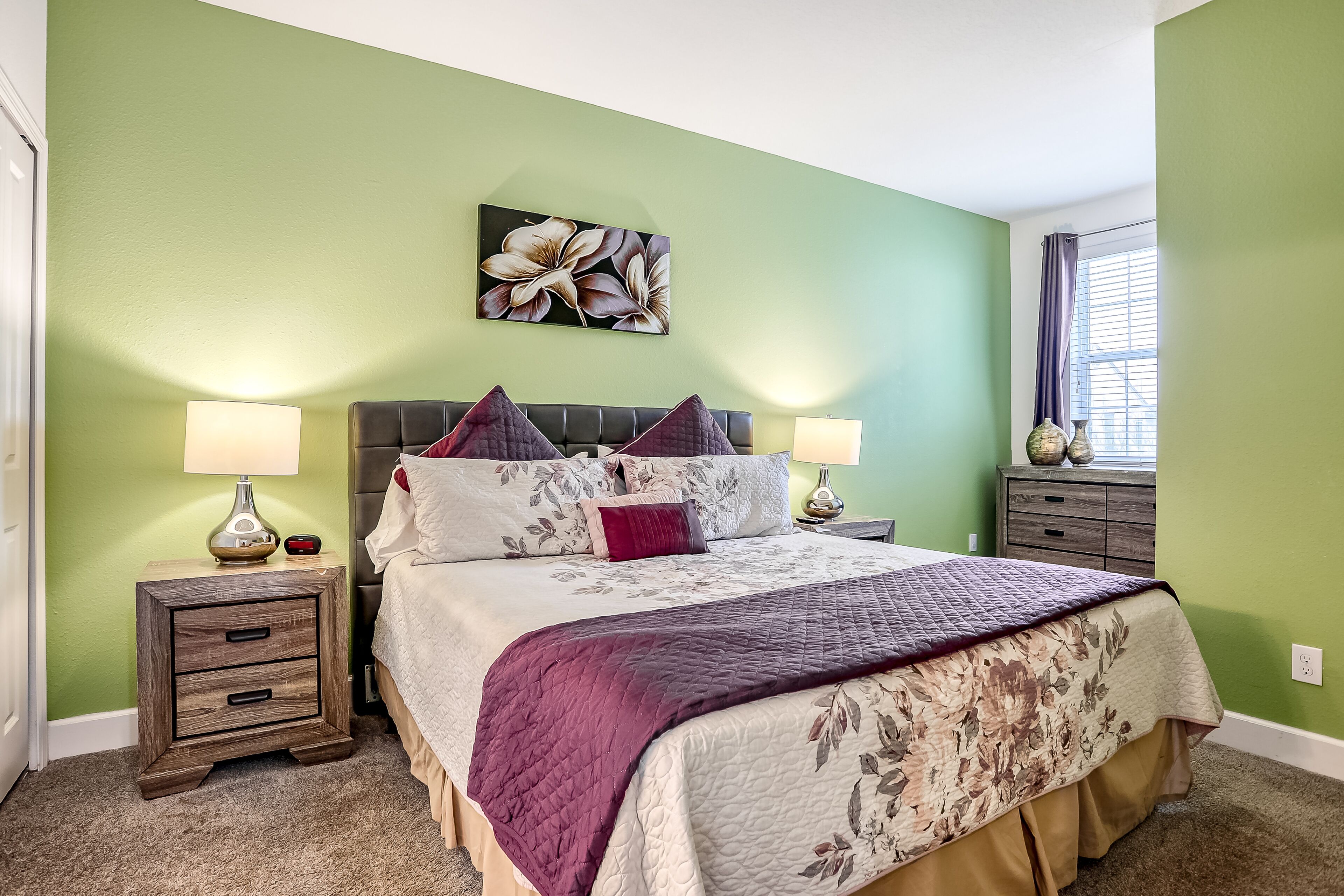 Une chambre à coucher comprenant un lit, deux tables de chevet, une fenêtre avec des rideaux et une décoration murale à motifs floraux.