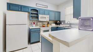 Una cocina con armarios azules, un refrigerador blanco, un microondas y un lavamanos. Hay un cartel en la pared y una ventana con persianas.