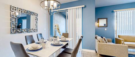 Un comedor moderno con una mesa blanca dispuesta para cuatro personas, rodeada de sillas oscuras. Un candelabro cuelga encima, y un espejo decorativo está en la pared.