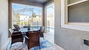 Uma área de varanda coberta com uma mesa de vidro e cadeiras de vime, com vista para uma piscina e edifícios residenciais.
