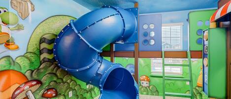 Zona de juegos infantiles con un tobogán azul, una pared de escalar verde y un mural con hongos y un personaje.