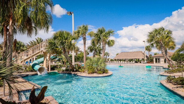 Area piscina di un resort con uno scivolo acquatico, palme e un edificio con tetto di paglia.
