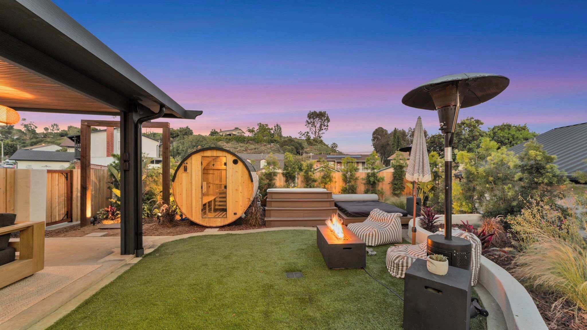 Twilight backyard spa zone — barrel sauna, hot tub, fire pit lounge, and patio heater under a pink-purple sky