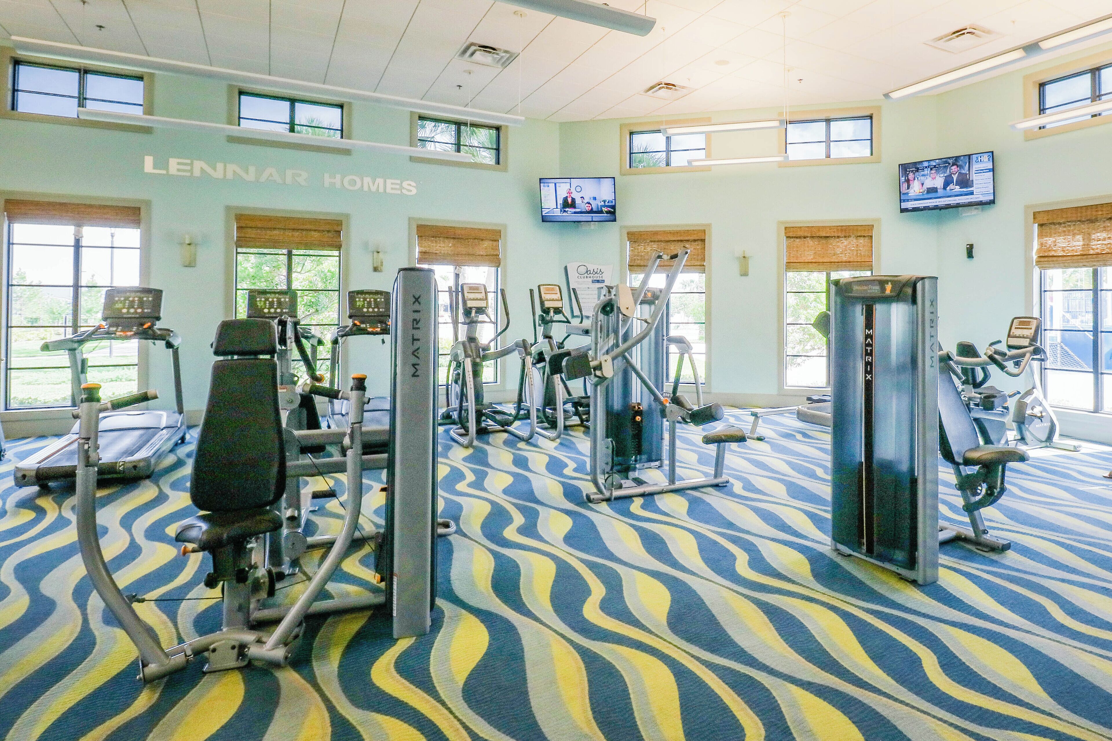 A gym with various exercise machines, large windows, and a patterned carpet.