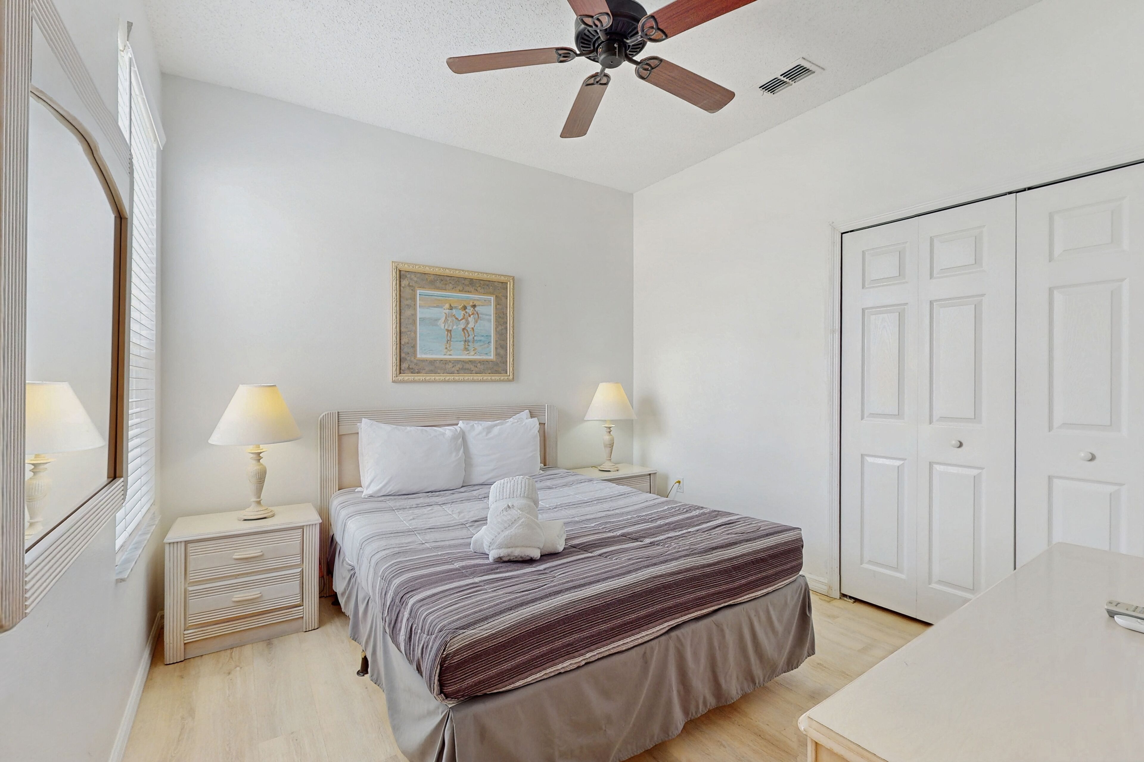 A bedroom with a bed, bedside lamps, a ceiling fan, a framed picture on the wall, and a wardrobe.