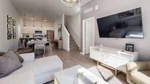 A modern living room with a white sofa, a coffee table, and a TV mounted on the wall. There is a staircase leading to the upper floor, and a kitchen area is visible in the background. - Two-story townhouse with private pool, screened lanai, washer/dryer (Davenport)