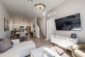 Ein modernes Wohnzimmer mit einem weißen Sofa, einem Couchtisch und einem an der Wand montierten Fernseher. Eine Treppe führt in die obere Etage, und im Hintergrund ist eine Küchenzeile erkennbar.