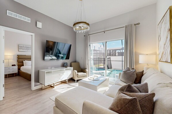 Ein modernes Wohnzimmer mit Sofa, Fernseher, Couchtisch und einer Glastür zum Poolbereich.