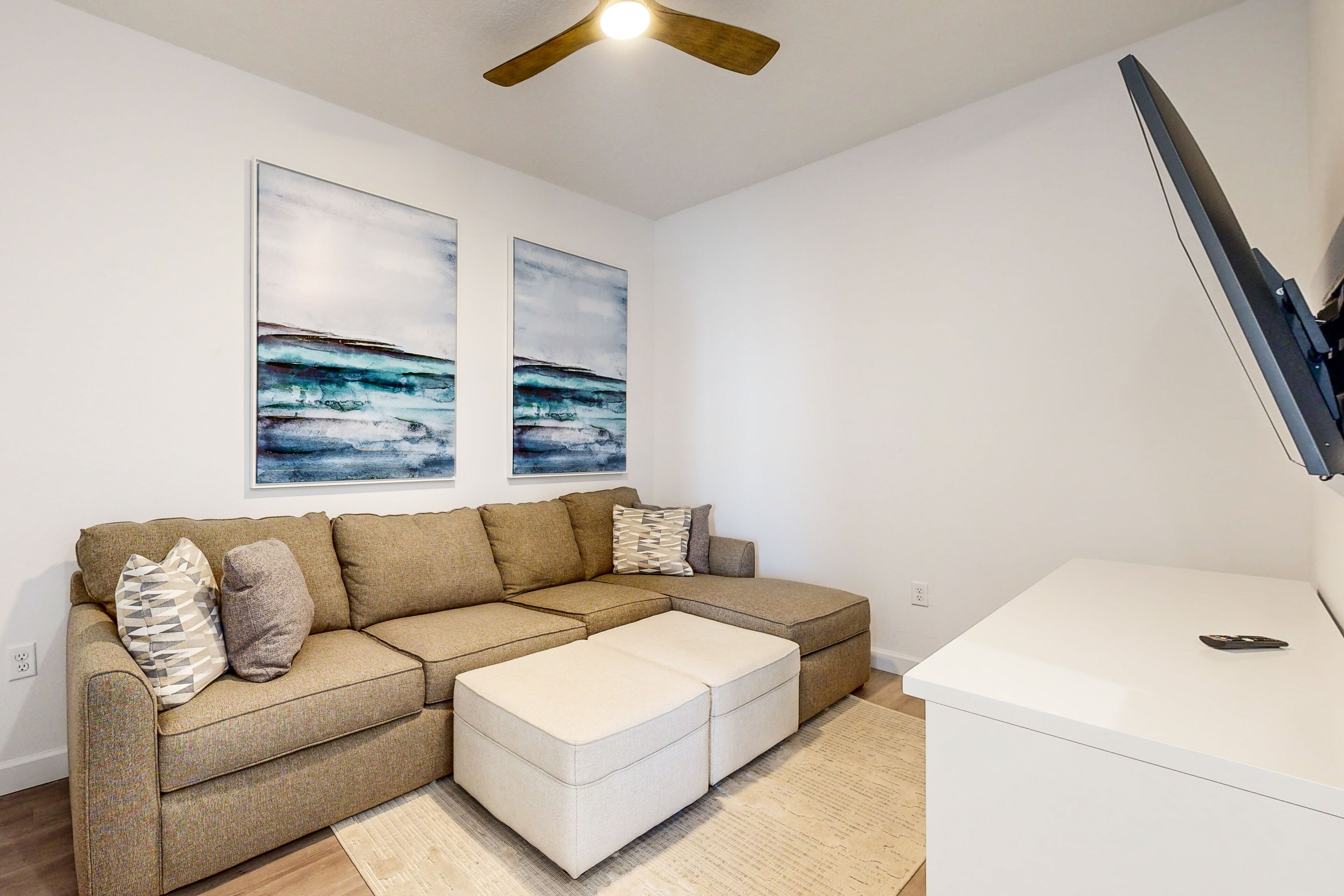 A modern living room with a sectional sofa, a white ottoman, a flat-screen TV mounted on the wall, and abstract artwork above the sofa.