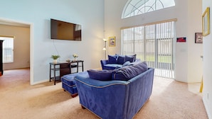 A living room with a blue sofa, a flat-screen TV, a small table with a plant, and a large window with a view of the outdoors. - Spacious home in cute area with central AC & washer/dryer - snowbirds welcome (Davenport)