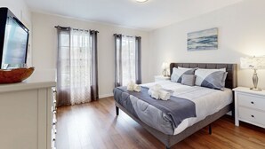 Een slaapkamer met een bed, nachtkastjes, gordijnen, een schilderij en een televisie op een dressoir.