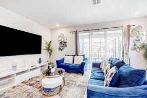 Een moderne woonkamer met een grote flatscreen-tv, blauwe banken, een witte tv-kast en een kamerbreed tapijt met dessin.