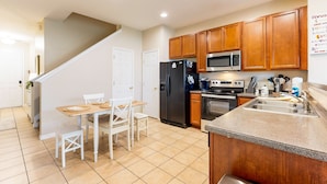 A kitchen with wooden cabinets, a black refrigerator, a microwave, a sink, and a dining area with a table and chairs. - Dog-friendly townhouse near Disney with outdoor pool, patio, streaming, W/D, AC (Kissimmee)