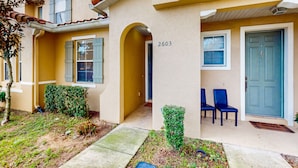 A two-story building with a beige facade, numbered 2603, featuring a small garden area with a pathway, two blue chairs, and a door with a blue handle. - Dog-friendly townhouse near Disney with outdoor pool, patio, streaming, W/D, AC (Kissimmee)