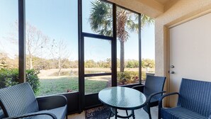 A covered patio with wicker furniture, a glass table, and a view of a landscaped area with trees and a palm tree. - Family Home w/Shared Pool,WiFi,AC,Lake Views - Close to Walt Disney World Resort (Kissimmee)