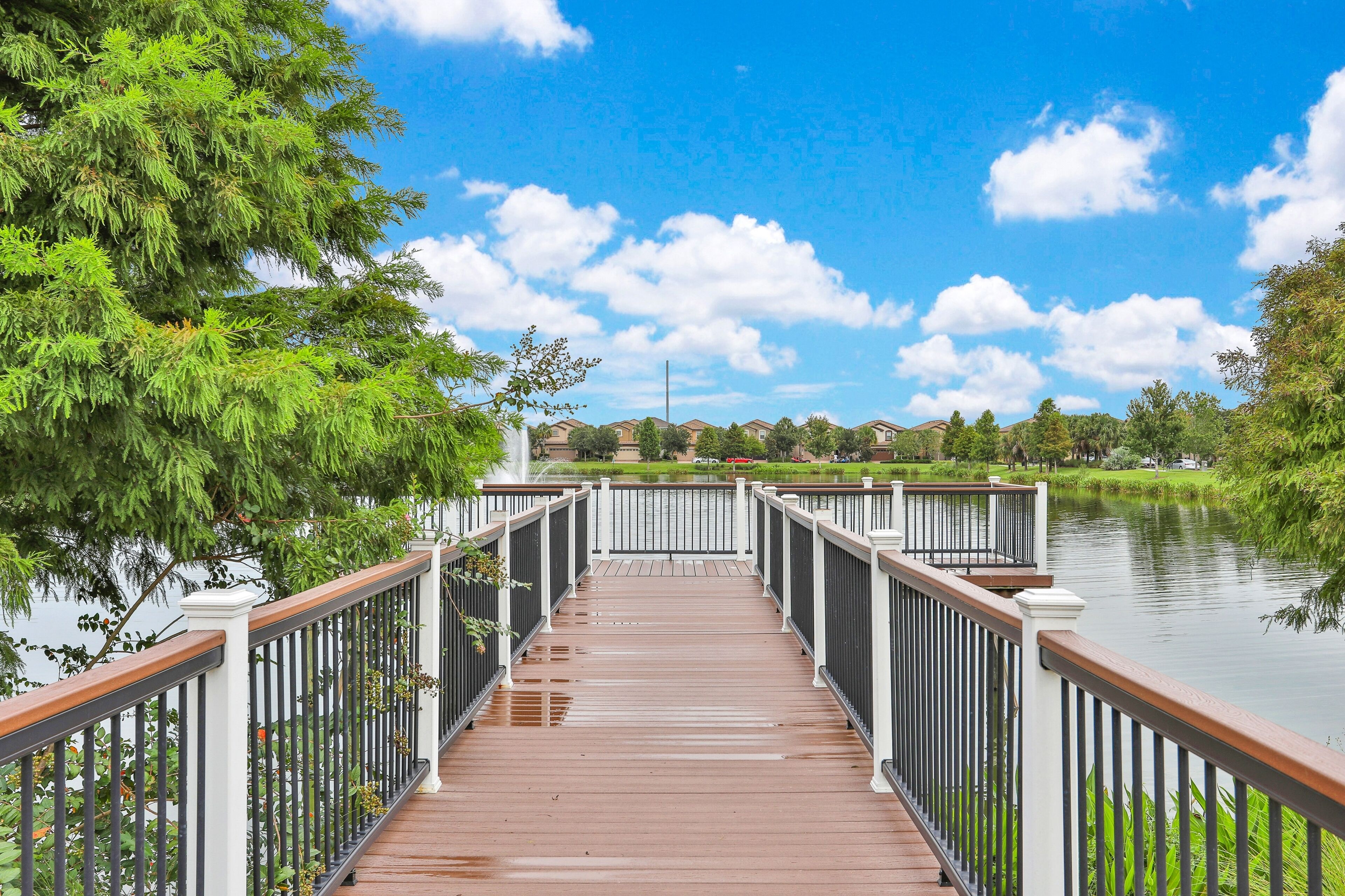 A wooden bridge with white railings leading to a body of water, surrounded by greenery and houses in the background.