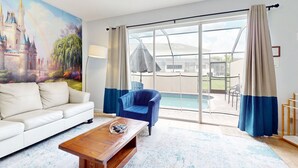 A room with a white sofa, a blue armchair, a wooden coffee table, and a large window with a view of a swimming pool and outdoor seating. - Resort home with private pool, hot tub, tennis, game room, gym, sports courts (Kissimmee)